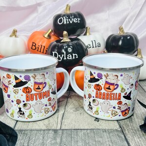 K&ouml;nnte beinhalten: Zwei wei&szlig;e Emailletassen mit Halloween-Motiven. Eine Tasse sagt "Autumn" und die andere sagt "Arabella". Die Tassen sind von kleinen K&uuml;rbissen mit darauf geschriebenen Namen umgeben.
