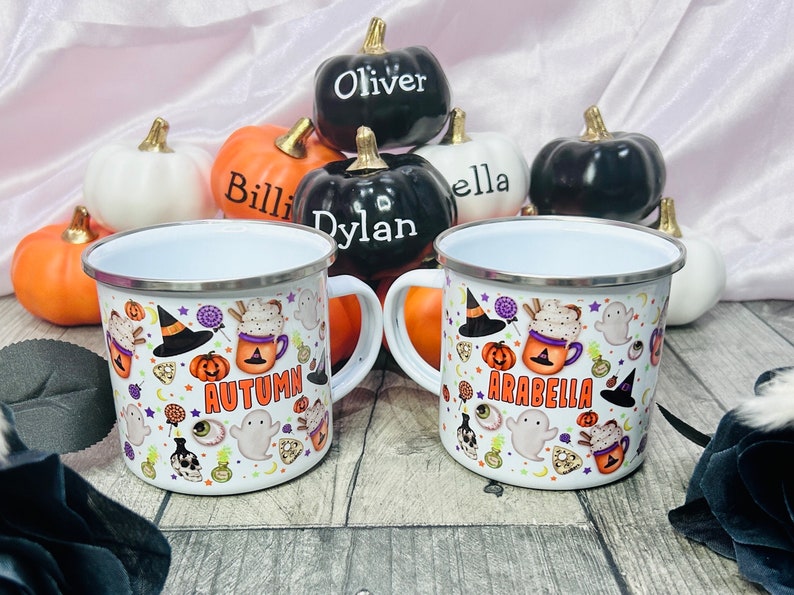 K&ouml;nnte beinhalten: Zwei wei&szlig;e Emailletassen mit Halloween-Motiven. Eine Tasse sagt "Autumn" und die andere sagt "Arabella". Die Tassen sind von kleinen K&uuml;rbissen mit darauf geschriebenen Namen umgeben.