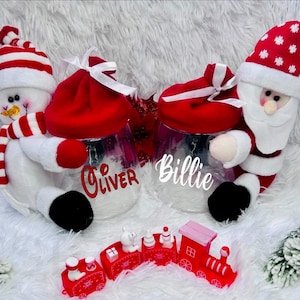 May include: Two clear glass jars decorated as a snowman and Santa Claus, each with a red lid and a name on the front. A small red toy train sits in front of the jars. The snowman jar has the name "Oliver" and the Santa jar has the name "Billie".