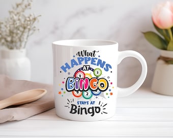 Op de afbeelding: Witte keramische koffiemok met een kleurrijk ontwerp waarop staat "What Happens at Bingo Stays at Bingo". Het ontwerp bevat een cirkel van bingoballen met nummers.