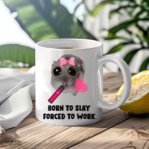 May include: A white ceramic mug featuring a graphic of a grey sugar glider with large black eyes, wearing a pink bow, and holding a pink vape pen that emits a pink cloud. The text BORN TO SLAY FORCED TO WORK is printed in black below the graphic.