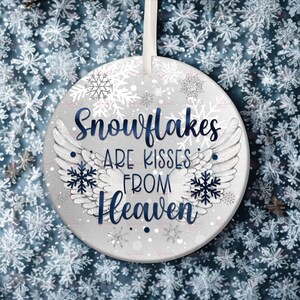 May include: A circular Christmas decoration in silver, inscribed with "Snowflakes are Kisses from Heaven" in navy blue. It features white angel wings and snowflake motifs, with a white ribbon for hanging.