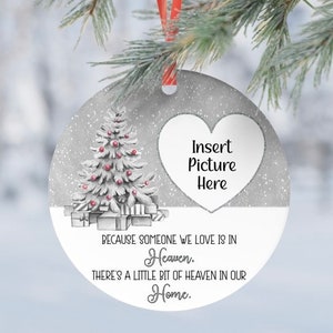 May include: A white and silver Christmas ornament with a white heart in the center. The heart has the text "Insert Picture Here". The ornament also has a white Christmas tree with red ornaments and a text that reads "Because someone we love is in Heaven, there's a little bit of Heaven in our Home."