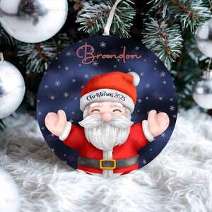 May include: Round Christmas ornament featuring Santa Claus in a red suit and hat, with a white beard and smiling face. The ornament has the name "Brandon" in red script and the text "Christmas 2025". The background is a dark blue with white snowflakes.
