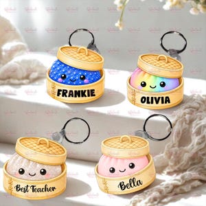 Puede incluir: Cuatro llaveros con personajes redondos y sonrientes dentro de cestas de vapor de bambú. Los personajes son azul con purpurina, arcoíris, blanco con purpurina y rosa sólido. Cada cesta está personalizada con los nombres Frankie, Olivia, Best Teacher y Bella.