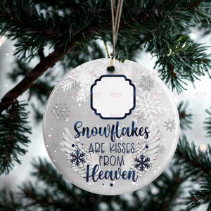 May include: A circular, grey Christmas bauble with the navy blue text "Snowflakes are kisses from Heaven". It features snowflake and wing designs, and a blank white space for a photo. The bauble hangs from a gold string.