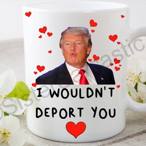 May include: White ceramic mug with a red heart and the text "I wouldn't deport you". A photo of Donald Trump blowing a kiss is in the background with red hearts.