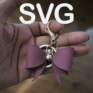 Könnte beinhalten: Ein goldener Schlüsselanhänger mit einem rosa Schleifenanhänger. Die Schleife ist aus Kunstleder und hat einen goldenen Metallring in der Mitte.