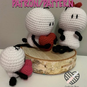 May include: Three crocheted amigurumi figures, each with a white body, black eyes, and black arms and legs. Two of the figures are holding red hearts, and the third figure is holding a pink heart. The figures are sitting on a wooden surface.