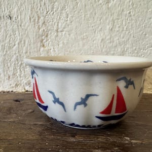 May include: A white ceramic bowl with a nautical design. The bowl features red sailboats and blue seagulls around the exterior. The bowl sits on a wooden surface.