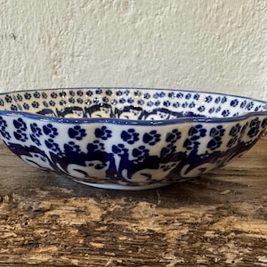 May include: A ceramic bowl with a scalloped edge, decorated with a blue and white floral pattern. The bowl has a rounded shape and is placed on a rustic wooden surface. The blue design includes stylized animal figures and floral motifs.