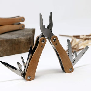 Könnte beinhalten: Ein Multitool mit Holzgriff ist geöffnet und zeigt eine Zange, eine Säge und andere Werkzeuge. Zwei weitere geschlossene Multitools liegen im Hintergrund auf einem Steinblock. Die Werkzeuge bestehen aus Edelstahl und Holz.