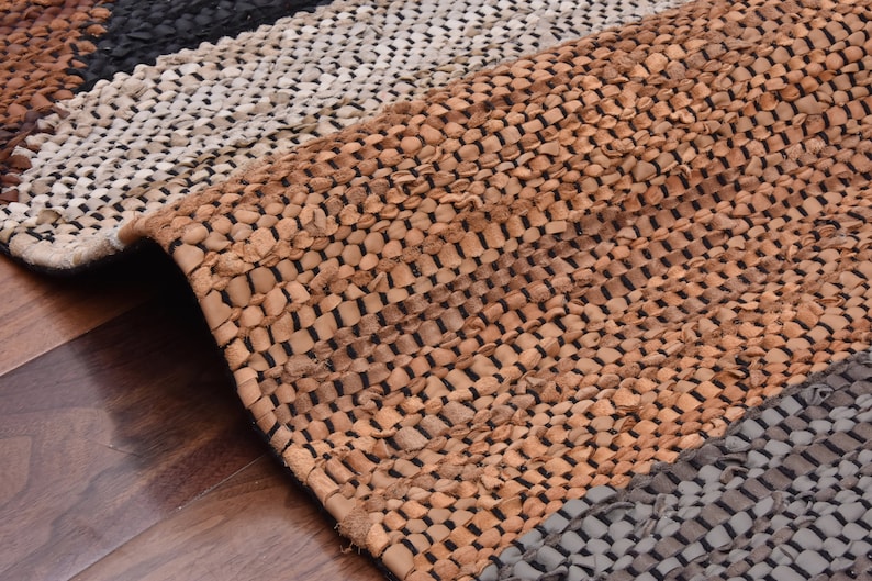 May include: A woven leather rug with a striped pattern in shades of brown, black, and white. The rug is made of braided leather strips and has a rustic, handmade look.