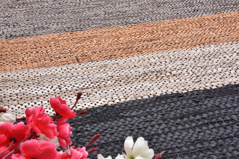 May include: A close-up of a multi-colored woven rug with a black, white, brown, and orange pattern. The rug is made of small, round pieces of material that are woven together. The rug is in focus, and the background is blurred.