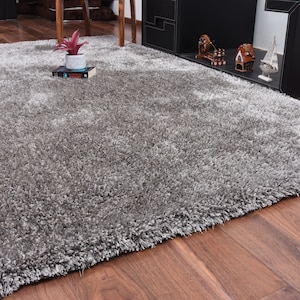 K&ouml;nnte beinhalten: Ein flauschiger, silbergrauer Teppich mit weicher, flauschiger Textur. Der Teppich liegt auf einem Holzboden.