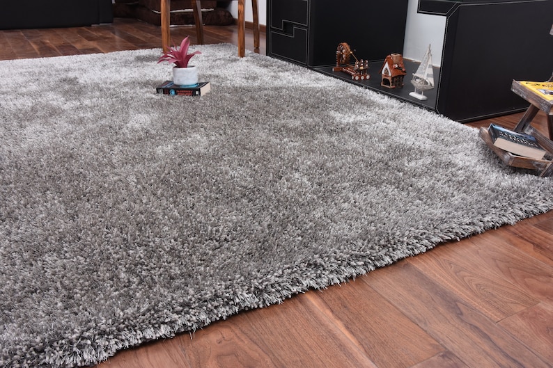 K&ouml;nnte beinhalten: Ein flauschiger, silbergrauer Teppich mit weicher, flauschiger Textur. Der Teppich liegt auf einem Holzboden.