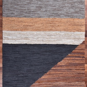 May include: A woven rug with a geometric design in shades of gray, brown, and beige. The rug has a textured surface and a modern, minimalist style.