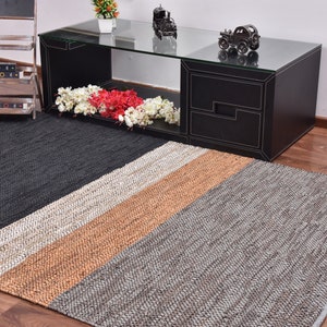 May include: A rectangular jute rug with a geometric pattern in shades of brown, gray, and white. The rug is placed on a hardwood floor in front of a black coffee table.