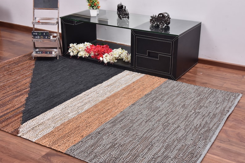 May include: A rectangular jute rug with a geometric pattern in shades of brown, gray, and white. The rug is placed on a hardwood floor in front of a black coffee table.