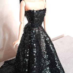 May include: A black strapless evening gown on a doll. The dress is covered in sequins and has a full skirt. The doll has pale skin and black hair. The dress appears to be handmade.