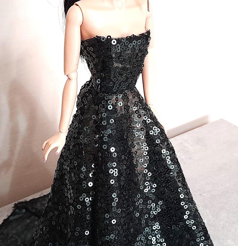 May include: A black strapless evening gown on a doll. The dress is covered in sequins and has a full skirt. The doll has pale skin and black hair. The dress appears to be handmade.