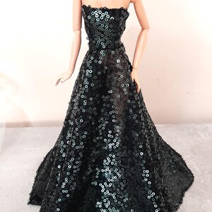 May include: A fashion doll wearing a strapless, floor-length gown. The dress is covered in black sequins, creating a shimmering effect. The doll has dark hair and is accessorized with long earrings.