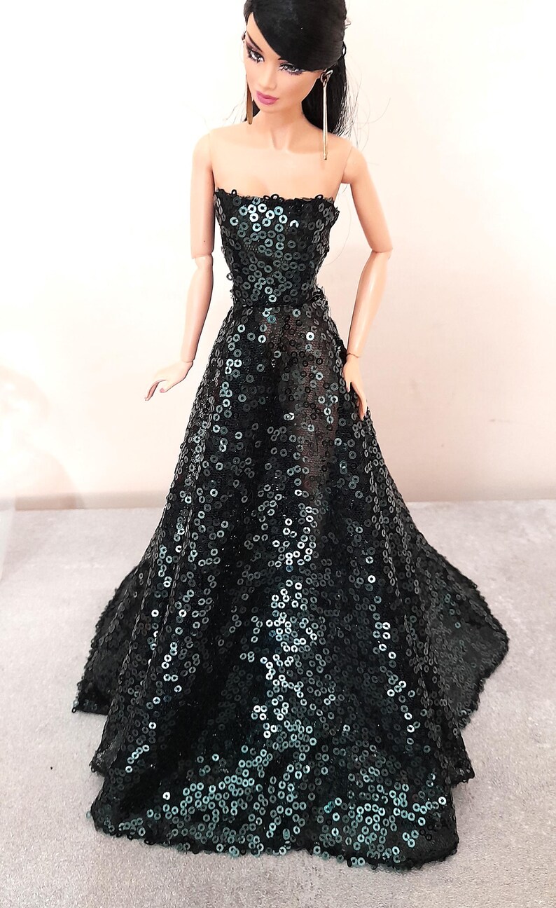 May include: A fashion doll wearing a strapless, floor-length gown. The dress is covered in black sequins, creating a shimmering effect. The doll has dark hair and is accessorized with long earrings.