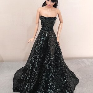 May include: A doll wearing a strapless, floor-length black gown. The dress is covered in sequins, creating a shimmering effect. The doll has dark hair and is posed in a standing position.