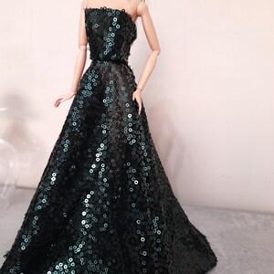 May include: A doll wearing a strapless, floor-length gown. The dress is covered in dark green sequins, creating a shimmering effect. The doll has long black hair and is wearing gold earrings.