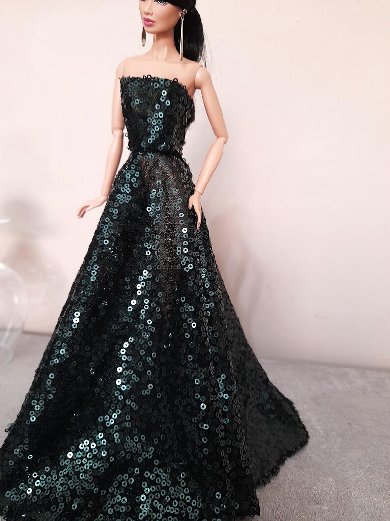 May include: A doll wearing a strapless, floor-length gown. The dress is covered in dark green sequins, creating a shimmering effect. The doll has long black hair and is wearing gold earrings.