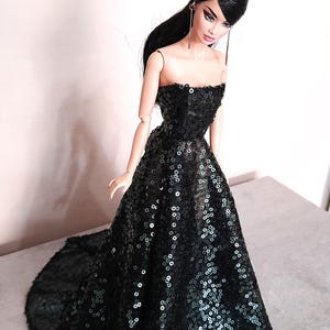 May include: A fashion doll wearing a strapless black sequin gown with a flowing skirt. The doll has long black hair and is wearing long, dangling earrings. The dress is covered in small, reflective sequins.