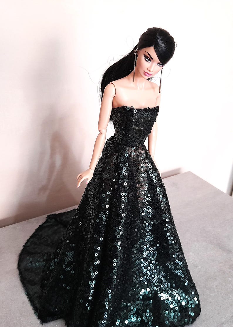 May include: A fashion doll wearing a strapless black sequin gown with a flowing skirt. The doll has long black hair and is wearing long, dangling earrings. The dress is covered in small, reflective sequins.