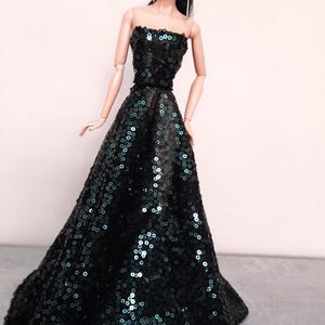 May include: A fashion doll wearing a strapless, floor-length gown. The dress is covered in dark green sequins, creating a shimmering effect. The doll has black hair and is wearing earrings.