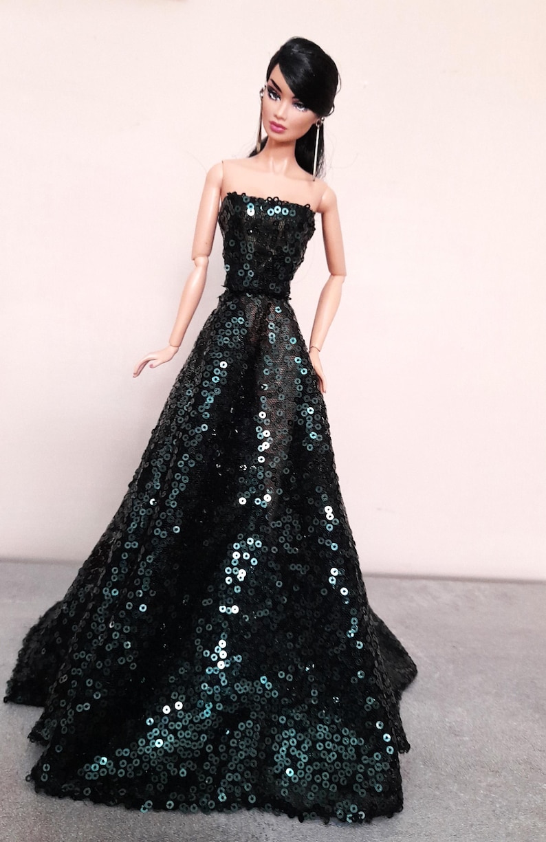 May include: A fashion doll wearing a strapless, floor-length gown. The dress is covered in dark green sequins, creating a shimmering effect. The doll has black hair and is wearing earrings.