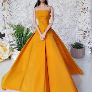 May include: A doll in a strapless, mustard-yellow gown with a full skirt. The doll has black hair and is positioned before a white lace backdrop. The dress has a fitted bodice and a flowing skirt, creating an elegant silhouette.