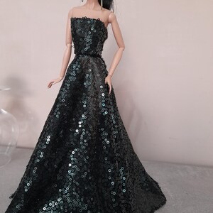 May include: A doll wearing a strapless, floor-length gown. The dress is dark green and covered in sequins. The doll has black hair pulled back in a ponytail and is wearing long earrings.