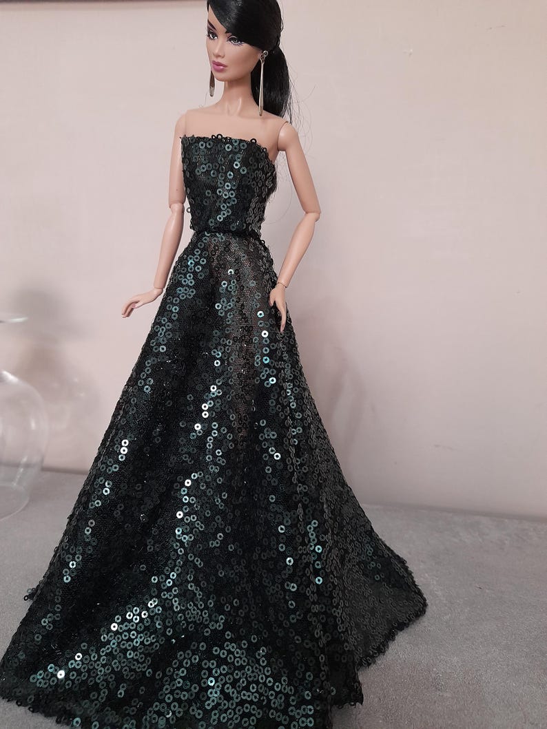 May include: A doll wearing a strapless, floor-length gown. The dress is dark green and covered in sequins. The doll has black hair pulled back in a ponytail and is wearing long earrings.