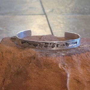 May include: A silver bracelet with an intricate design of mountains and other natural elements. The bracelet is made of sterling silver and has a polished finish.