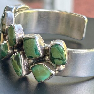 May include: A silver cuff bracelet with a row of green turquoise stones set in the metal. The stones are irregularly shaped and have a mottled pattern.