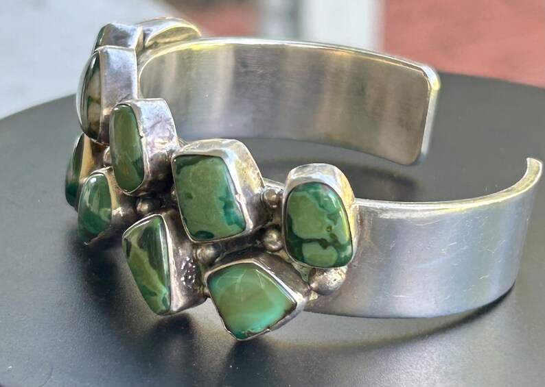 May include: A silver cuff bracelet with a row of green turquoise stones set in the metal. The stones are irregularly shaped and have a mottled pattern.