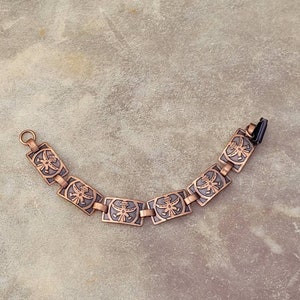 May include: A copper bracelet with a repeating design of stylized birds. The bracelet is made of linked rectangular pieces with a dark brown patina.