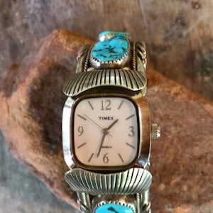 May include: A silver-toned Timex watch with a square face and turquoise accents. The watch features a white dial with black numerals and hands. The band is adorned with turquoise stones and decorative metalwork.