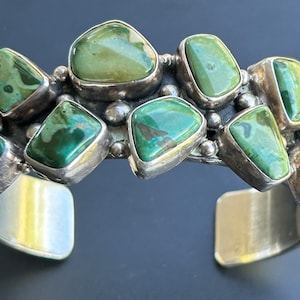 May include: A silver cuff bracelet with a row of green turquoise stones set in silver. The stones are irregularly shaped and have a mottled green color.