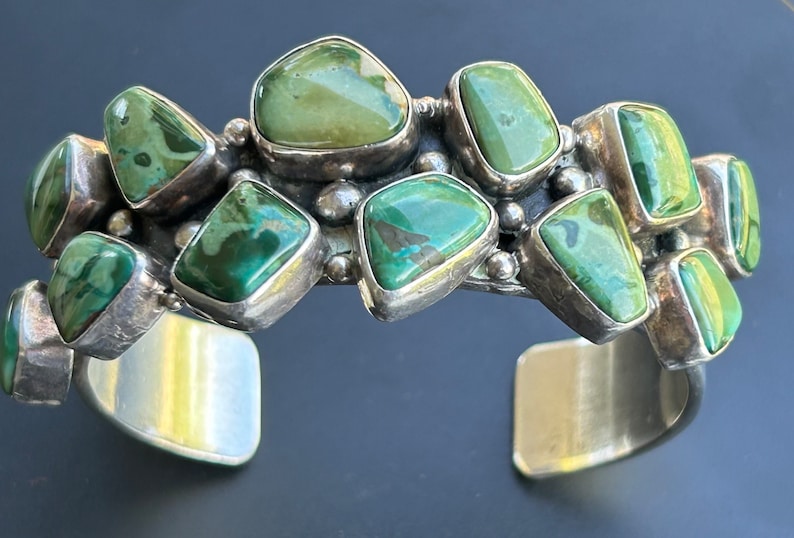 May include: A silver cuff bracelet with a row of green turquoise stones set in silver. The stones are irregularly shaped and have a mottled green color.