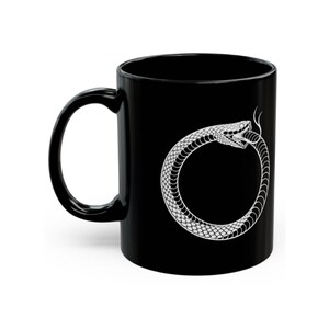 May include: Black ceramic mug featuring a white Ouroboros design, a snake eating its own tail. The handle is on the left side. The design is a detailed illustration of the snake's head and body forming a circle.