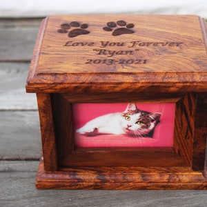 K&ouml;nnte beinhalten: Eine h&ouml;lzerne Tierurne mit einem Bilderrahmen und einer eingravierten Inschrift: "Love You Forever "Ryan" 2013-2021". Die Urne hat zwei eingravierte Pfotenabdr&uuml;cke auf der Oberseite.