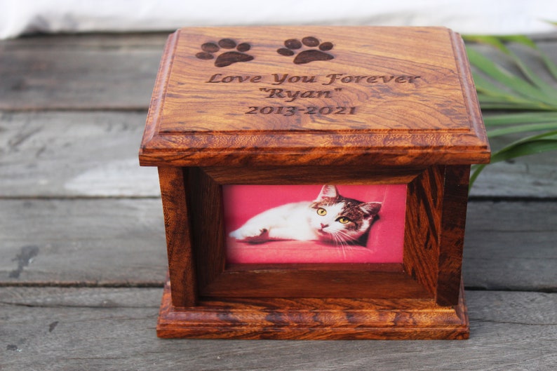K&ouml;nnte beinhalten: Eine h&ouml;lzerne Tierurne mit einem Bilderrahmen und einer eingravierten Inschrift: "Love You Forever "Ryan" 2013-2021". Die Urne hat zwei eingravierte Pfotenabdr&uuml;cke auf der Oberseite.