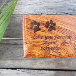 K&ouml;nnte beinhalten: Eine h&ouml;lzerne Tierurne mit einem Pfotenabdruck-Design. Die Urne ist mit dem Text "Love You Forever "Ryan" 2013-2021" graviert.