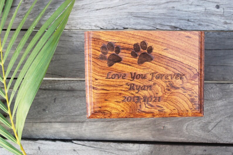 K&ouml;nnte beinhalten: Eine h&ouml;lzerne Tierurne mit einem Pfotenabdruck-Design. Die Urne ist mit dem Text "Love You Forever "Ryan" 2013-2021" graviert.