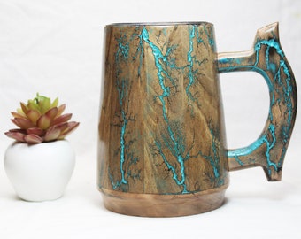 Può includere: Una tazza di legno con un design marmoreo blu turchese e marrone. La tazza ha un manico grande e una base larga.
